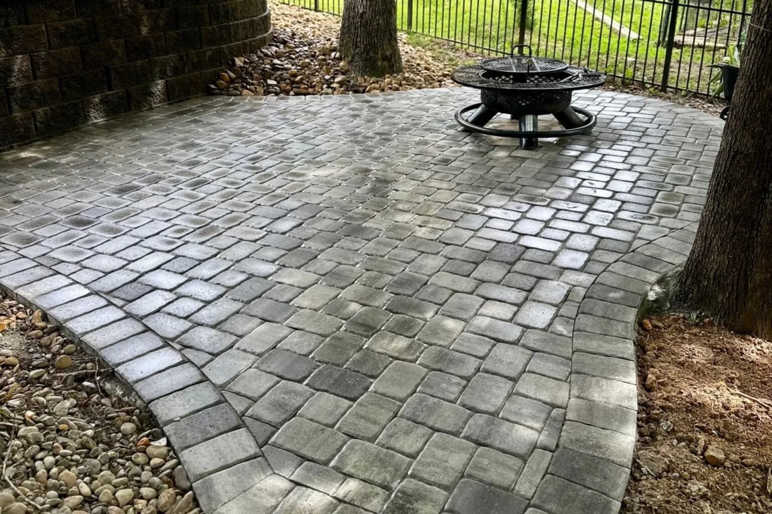 Backyard Escape shaded paver patio with a fire pit beneath mature trees and a curved retaining wall
