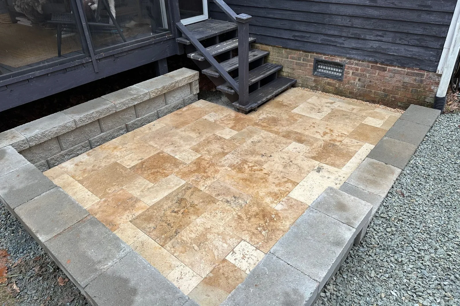 Mixed-tone travertine patio bounded by a capped block wall below existing wooden porch stairs
