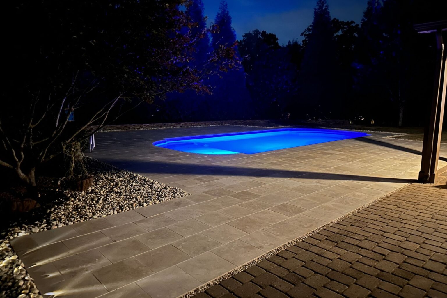 Night view across the illuminated pool, paver patio, and rock border