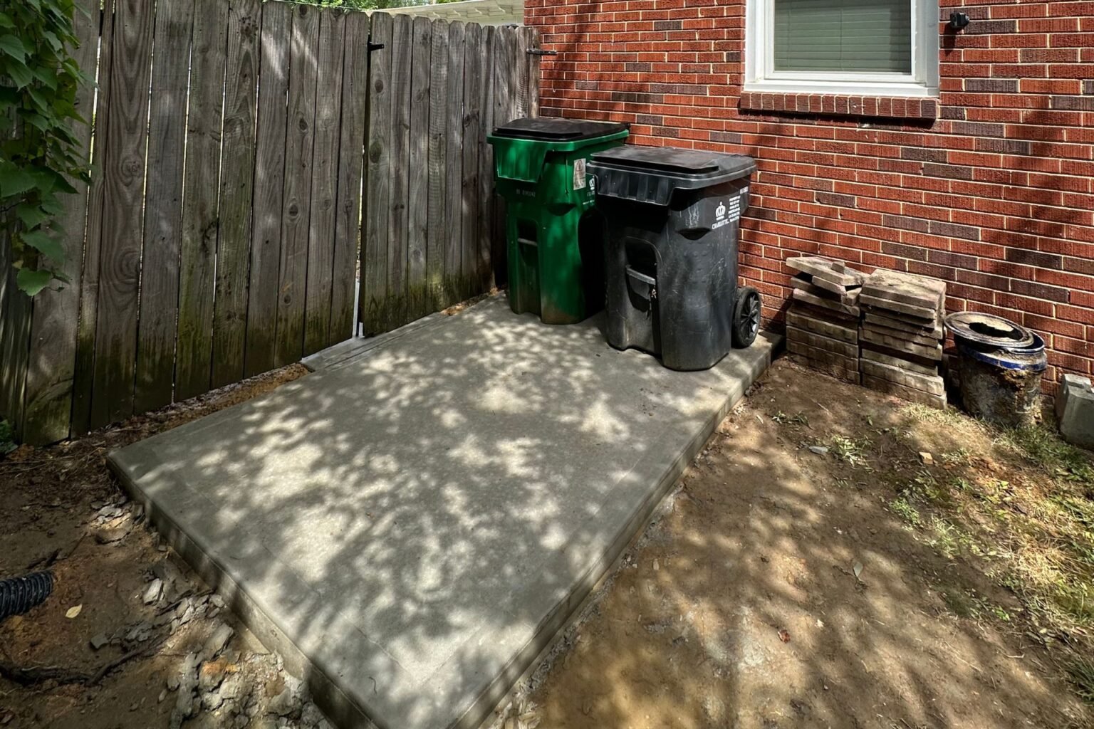Finished rectangular concrete pad holding trash bins beside the brick home and wood gate