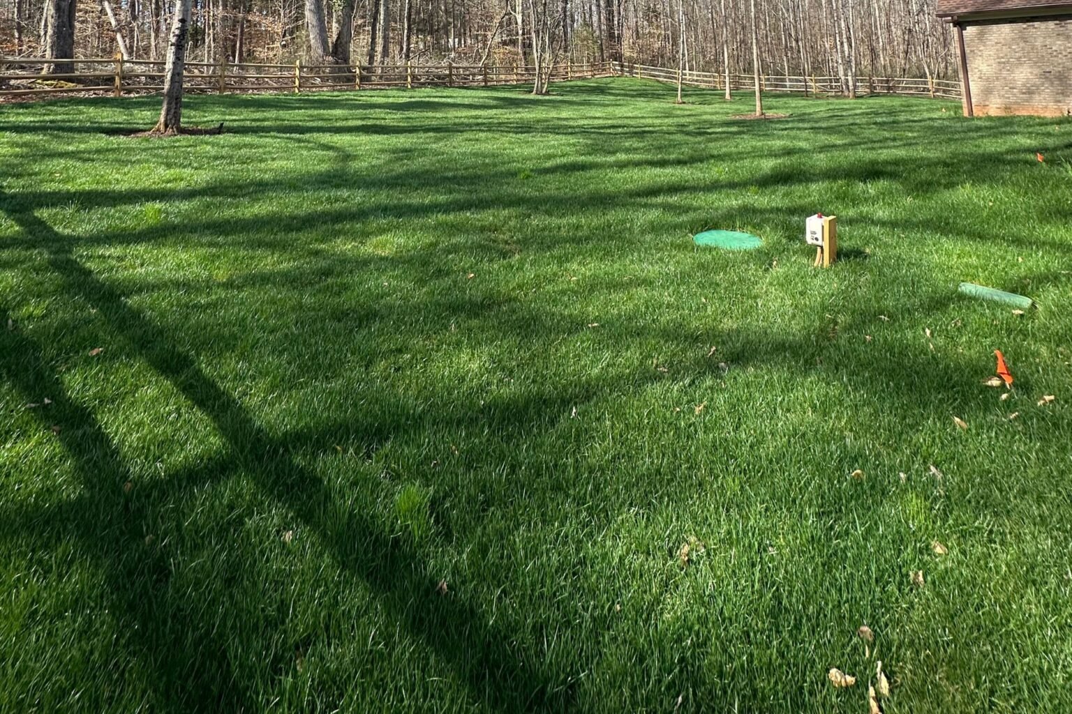Green backyard lawn with utility-style covers, marker flags, fencing, and woods