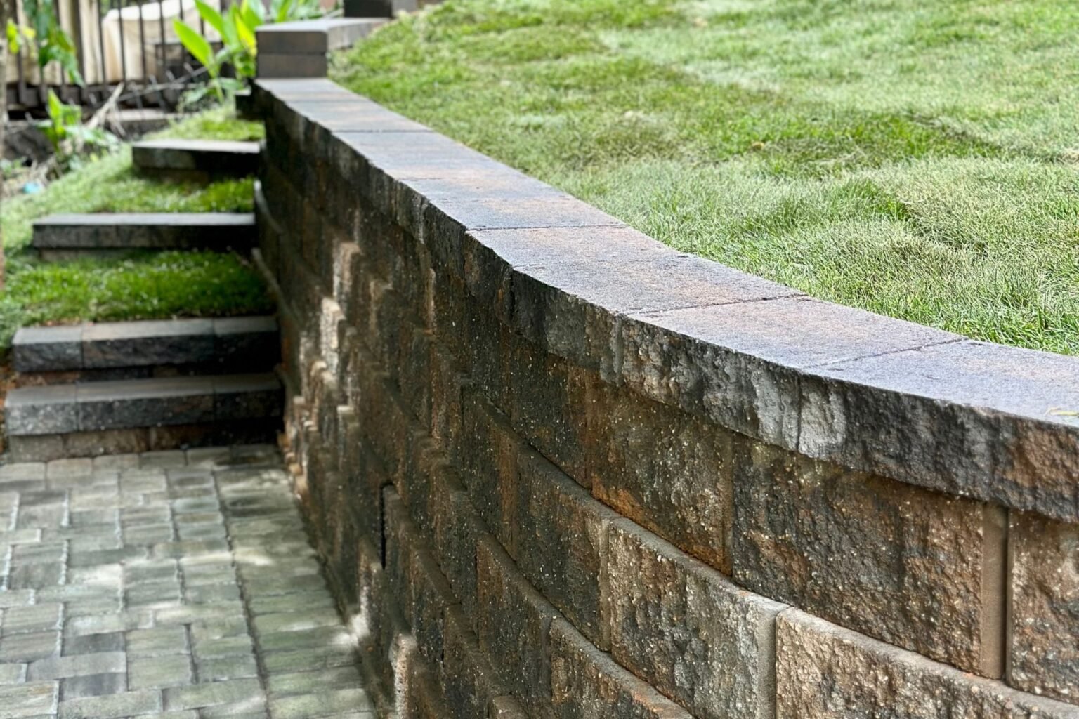Paver patio below a curved block retaining wall, with green sod and masonry steps